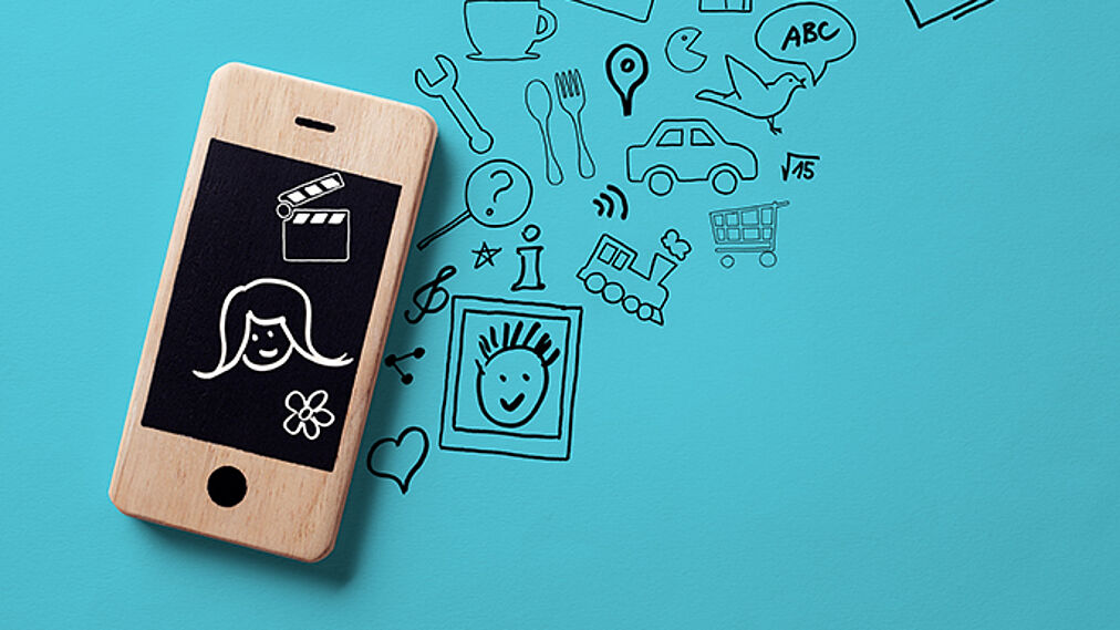 Das Foto zeigt ein Modell eines Smartphones aus Holz auf einem türkisen Hintergrund. Auf dem Handybildschirm und - wie daraus wachsend - darüber hinaus auf dem Hintergrund sind Symbole skizziert, die für die Lebenswelt von Jugendlichen stehen: Gesichter, Blumen, Videoklappe, ein Herz, Messer und Gabel, eine Tasse, ein Einkaufswagen, das WLAN-Zeichen, eine Filmklappe und andere