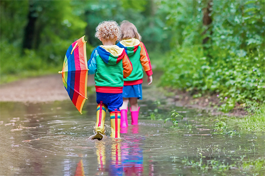 La photo montre deux enfants de dos qui marchent dans une grande gouille. Ils portent des habits et des bottes de pluie multicolores. Un enfant tient un parapluie aux couleurs de l’arc-en-ciel. 